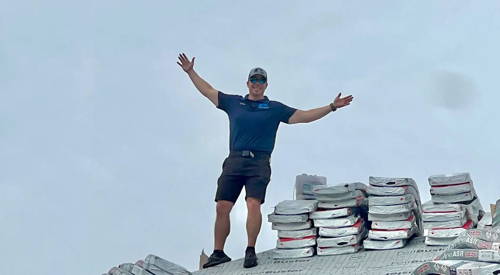 Joshua Holmes standing on a completed roof with shingle bundles staged nearby, arms raised