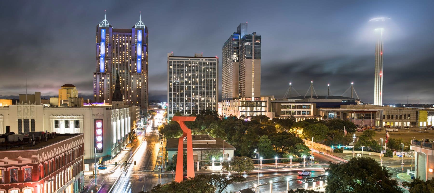 A vibrant nighttime cityscape featuring tall buildings, bright lights, and a prominent red sculpture in the foreground