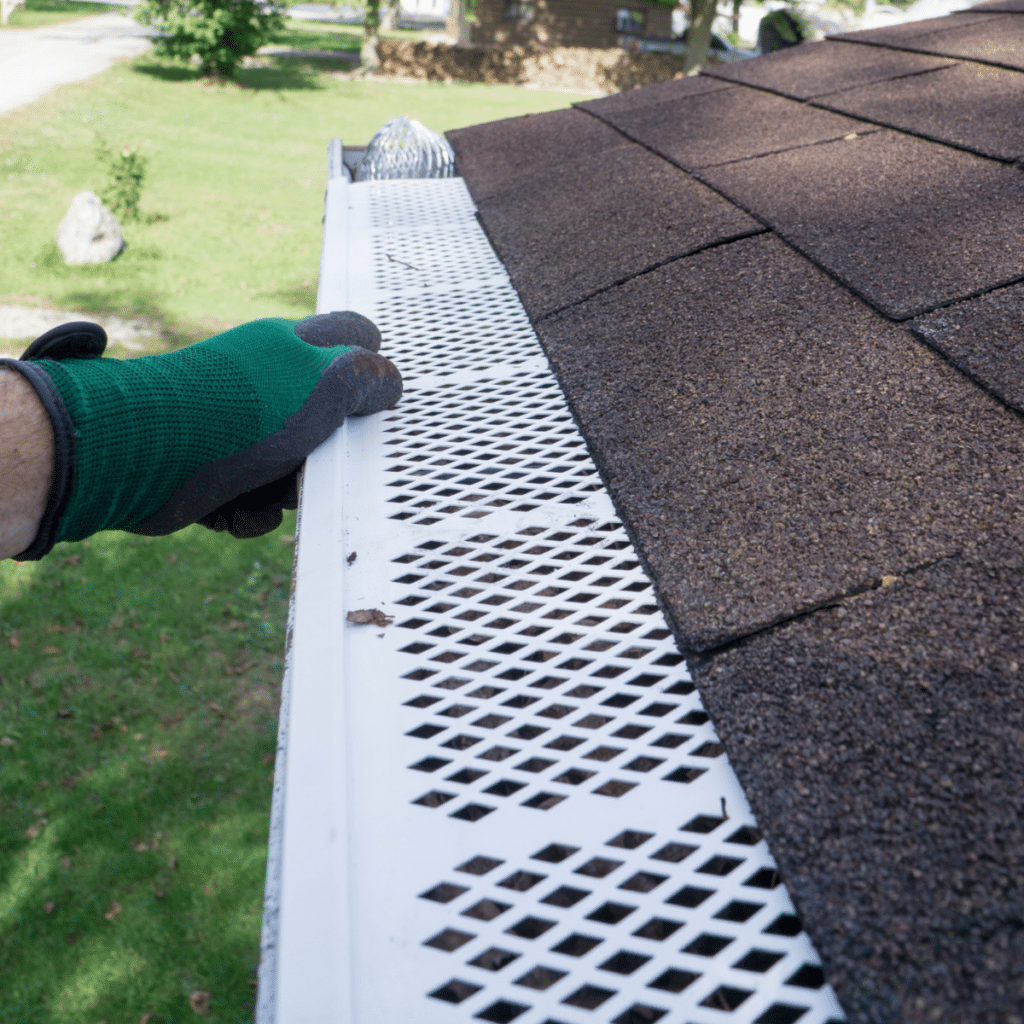 A gloved hand adjusting a white gutter guard on a roof to prevent debris accumulation