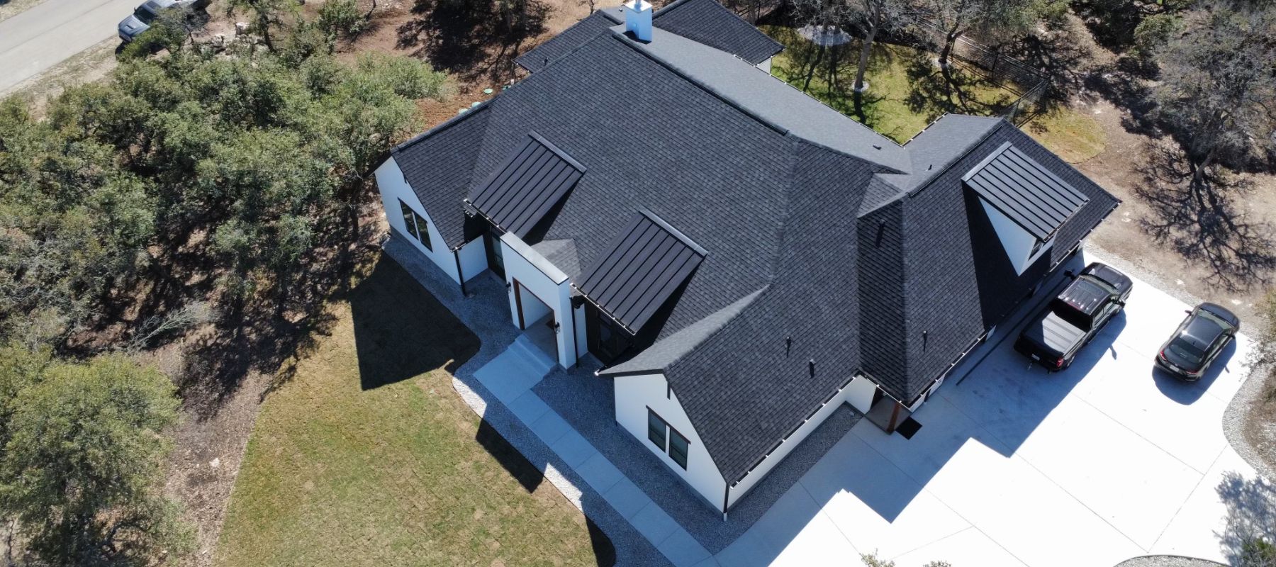 Aerial view of a modern house with a black shingle roof surrounded by trees and a clean driveway