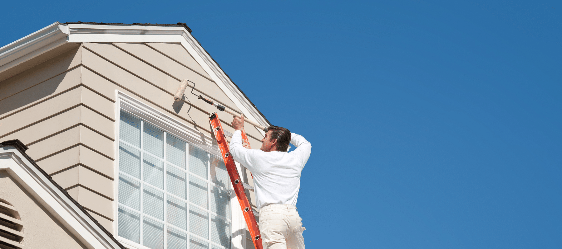 professional house painter standing on a ladder using a long roller to roll on paint to a home exterior