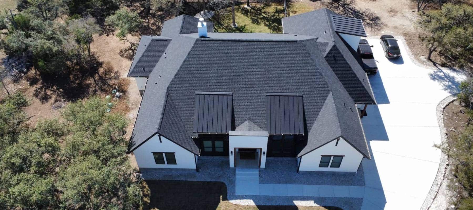 Aerial view of a modern house with a black shingle roof surrounded by trees and a clean driveway