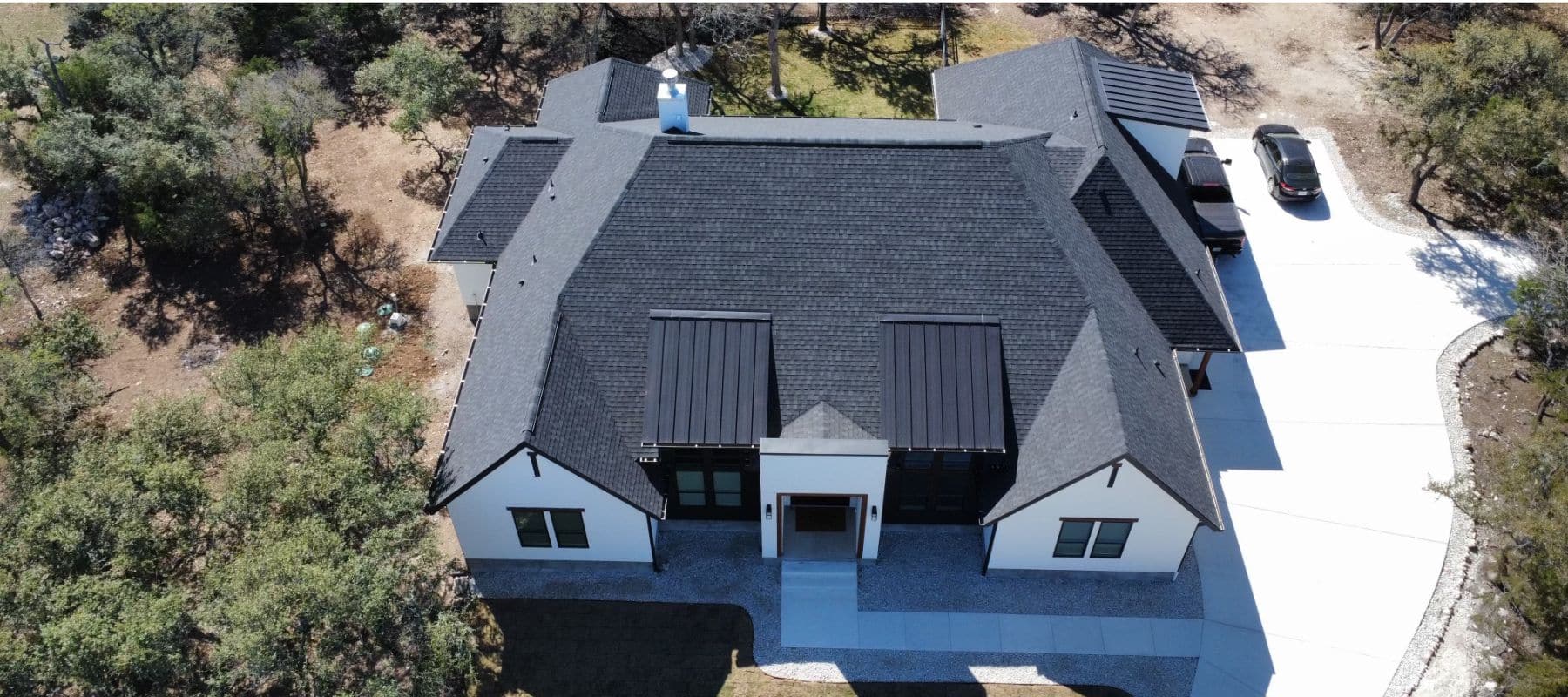 Aerial view of a modern house with a black shingle roof surrounded by trees and a clean driveway