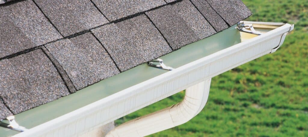 A clean gutter system attached to a shingle roof, with a downspout leading to a green lawn below
